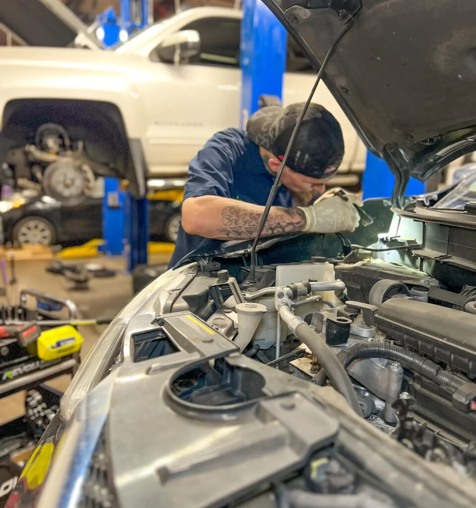 A mechanic works under the hood of a car surrounded by tools with a lifted vehicle in the background at an auto repair shop e1740500276643 ezgif.com webp to jpg converter
