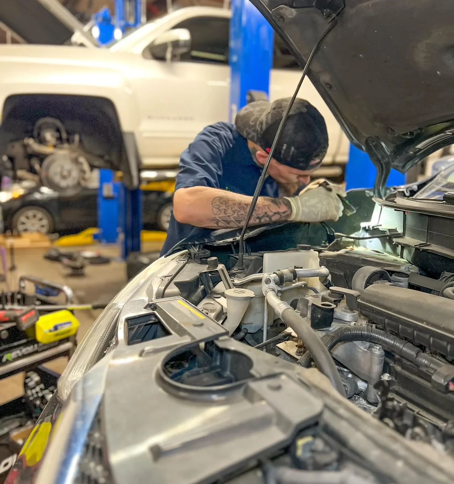 A mechanic works under the hood of a car surrounded by tools with a lifted vehicle in the background at an auto repair shop e1740500276643 ezgif.com webp to jpg converter